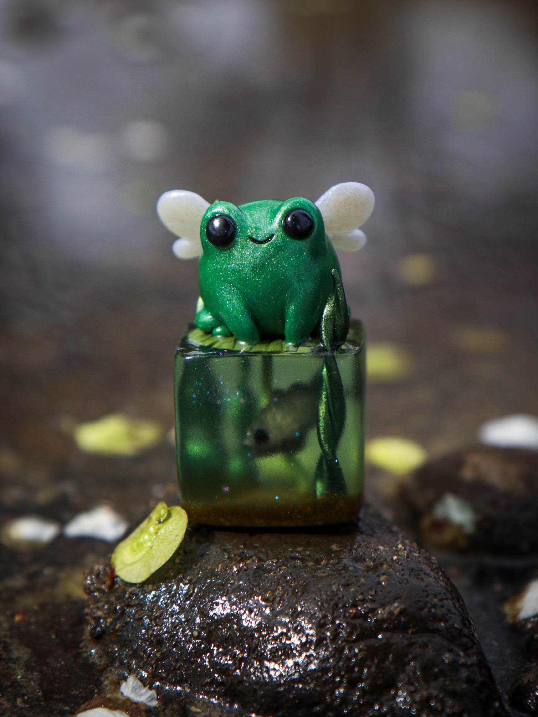 A small green frog figurine with wings on a cube, part of the Little Shit Big Deal - CUBES collection by Fairies And Fancies. Sculpted by Dr. Polpz. Ships randomly.