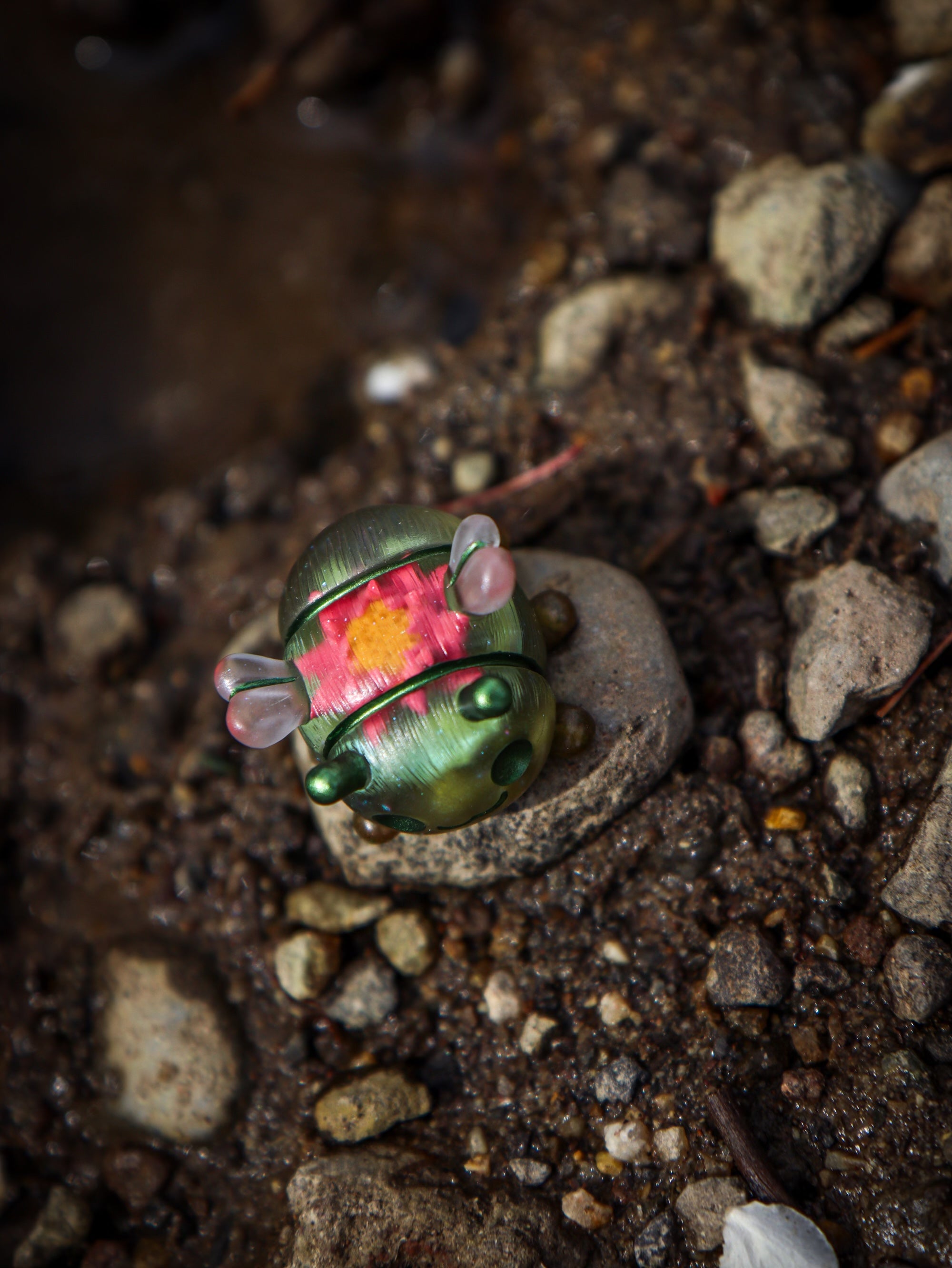 A small green and pink beetle toy on a rock, part of the Octopus Cube collection by Fairies And Fancies at Strangecat Toys. Made of Polymer Clay and Resin.