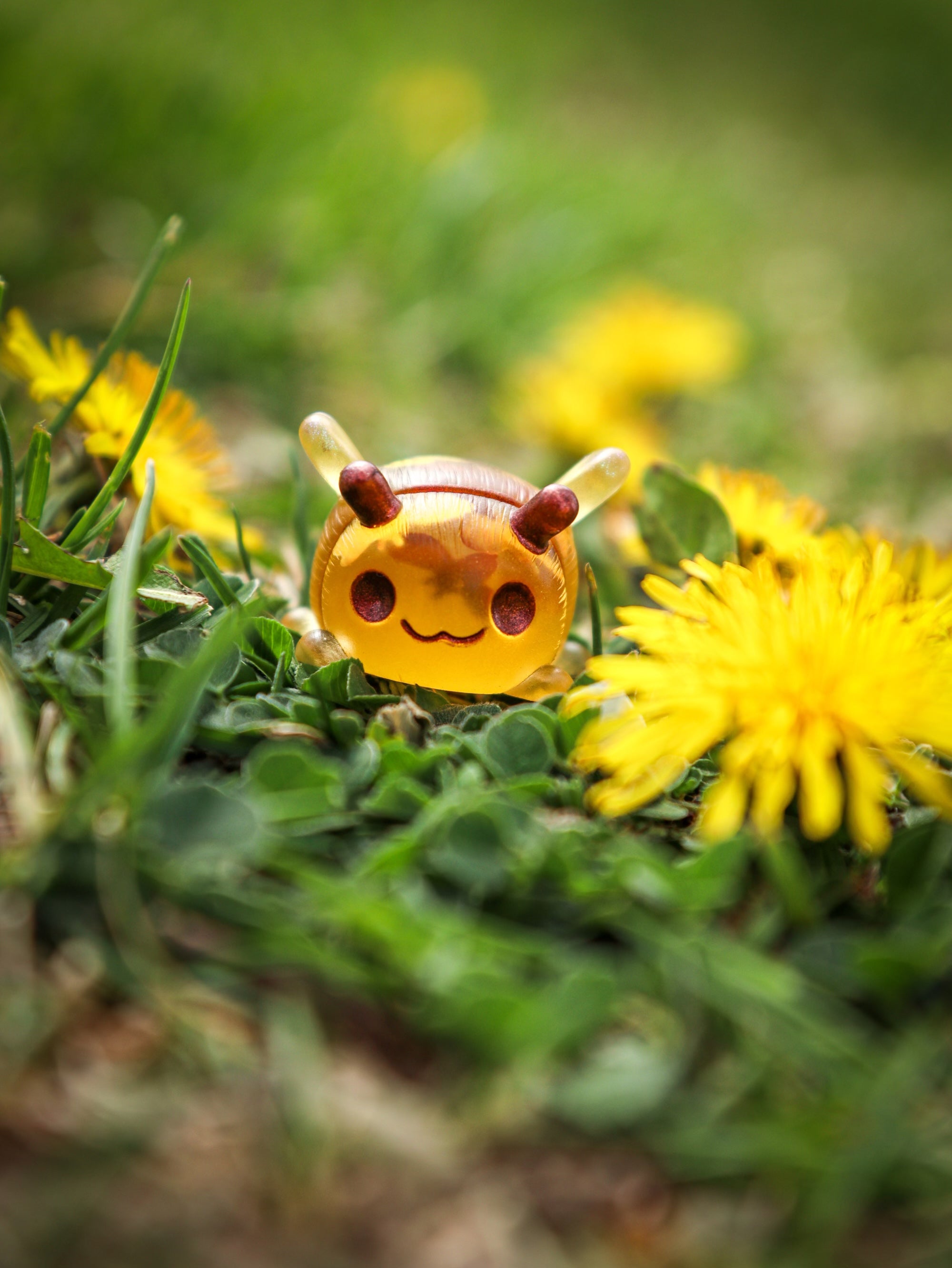 A yellow insect toy on grass with flowers, part of the GRUBLETS collection by Fairies And Fancies at Strangecat Toys. Approximately 1-2 inches tall, made of Polymer Clay and Resin.