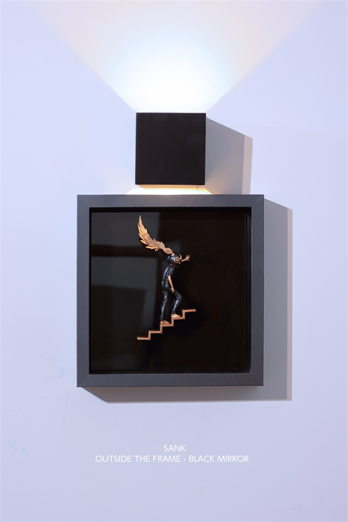 Sank - Outside the Frame: A minimalist wall-mounted art piece featuring a tiny black figurine with one golden wing, in a shadow box beneath a cube lamp.