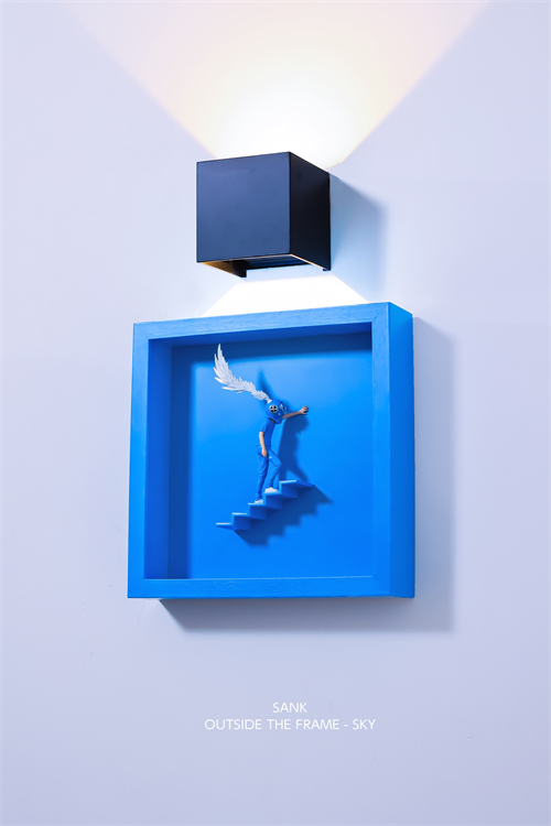 Sank - Outside the Frame art piece with blue figurine, steps, and feathered wing, mounted under a black sconce. Preorder, ships end of Q4 2025.