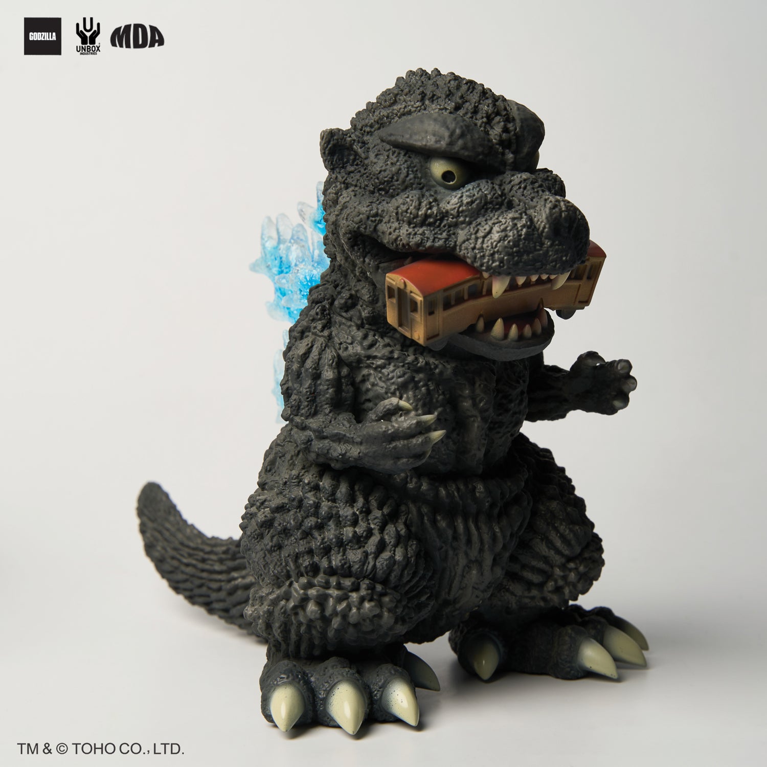 GODZILLA 1954 MDA PROJECT - Preorder: Stylized vinyl figure of Godzilla biting a toy train, featuring charcoal texture and blue dorsal plates.
