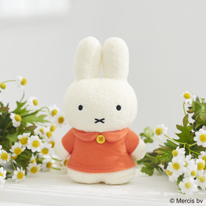 Tokotoko Club Miffy plush toy standing in an orange coat with a yellow button, surrounded by white flowers, embodying the playful art toy spirit.