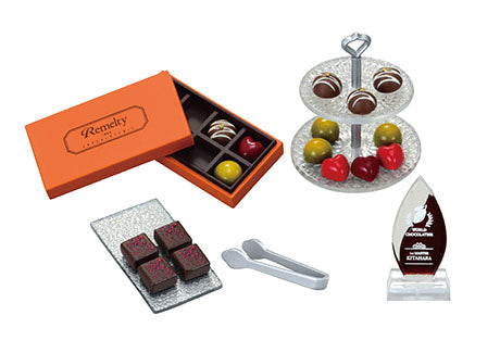 Display of gourmet chocolates featuring the Chocolaterie Re-ment Blind Box Series with an orange gift box, bonbons, dark chocolate squares, and a clear award plaque.