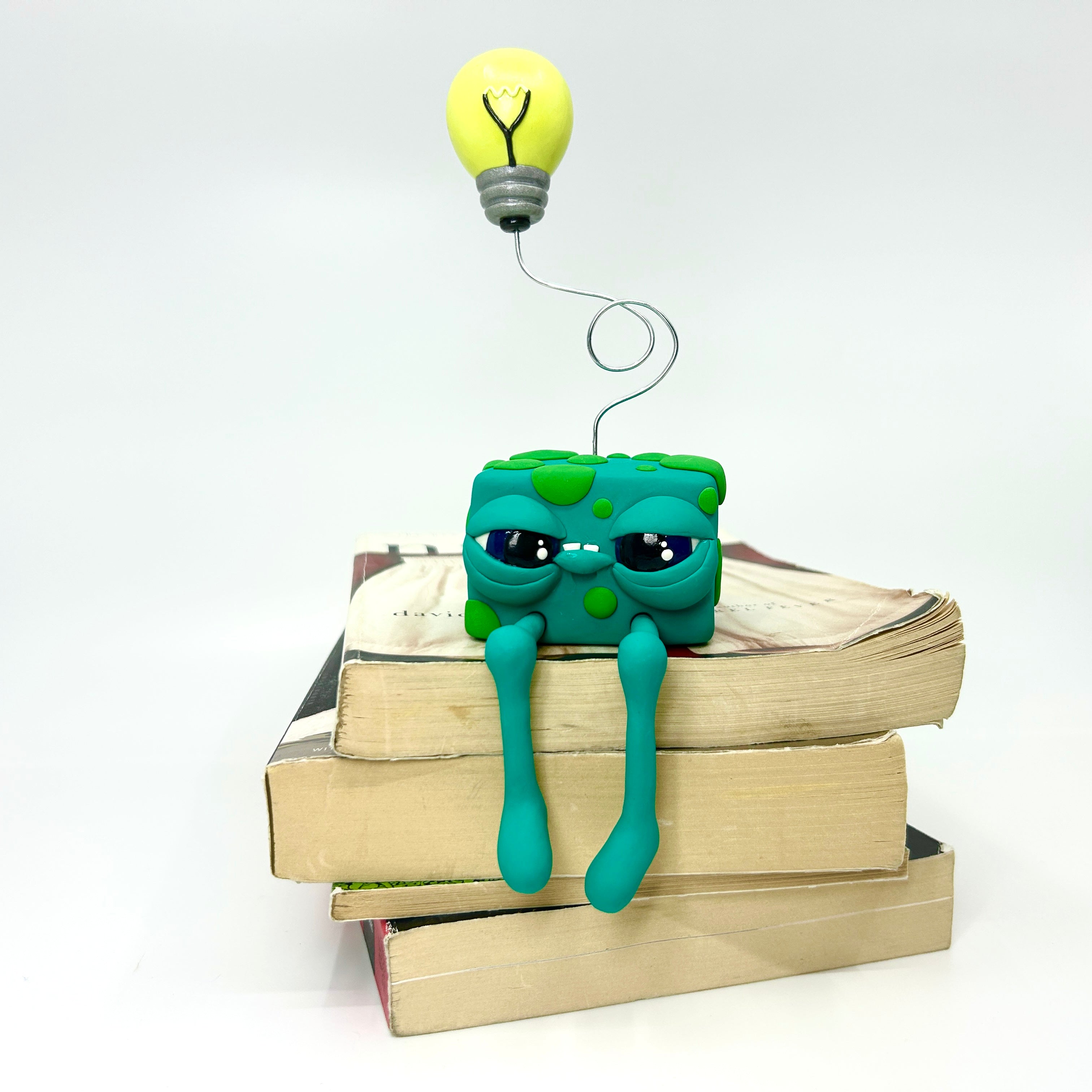 Toy on books, green plasticine creature, yellow light bulb, blue balloon, wire close-up.