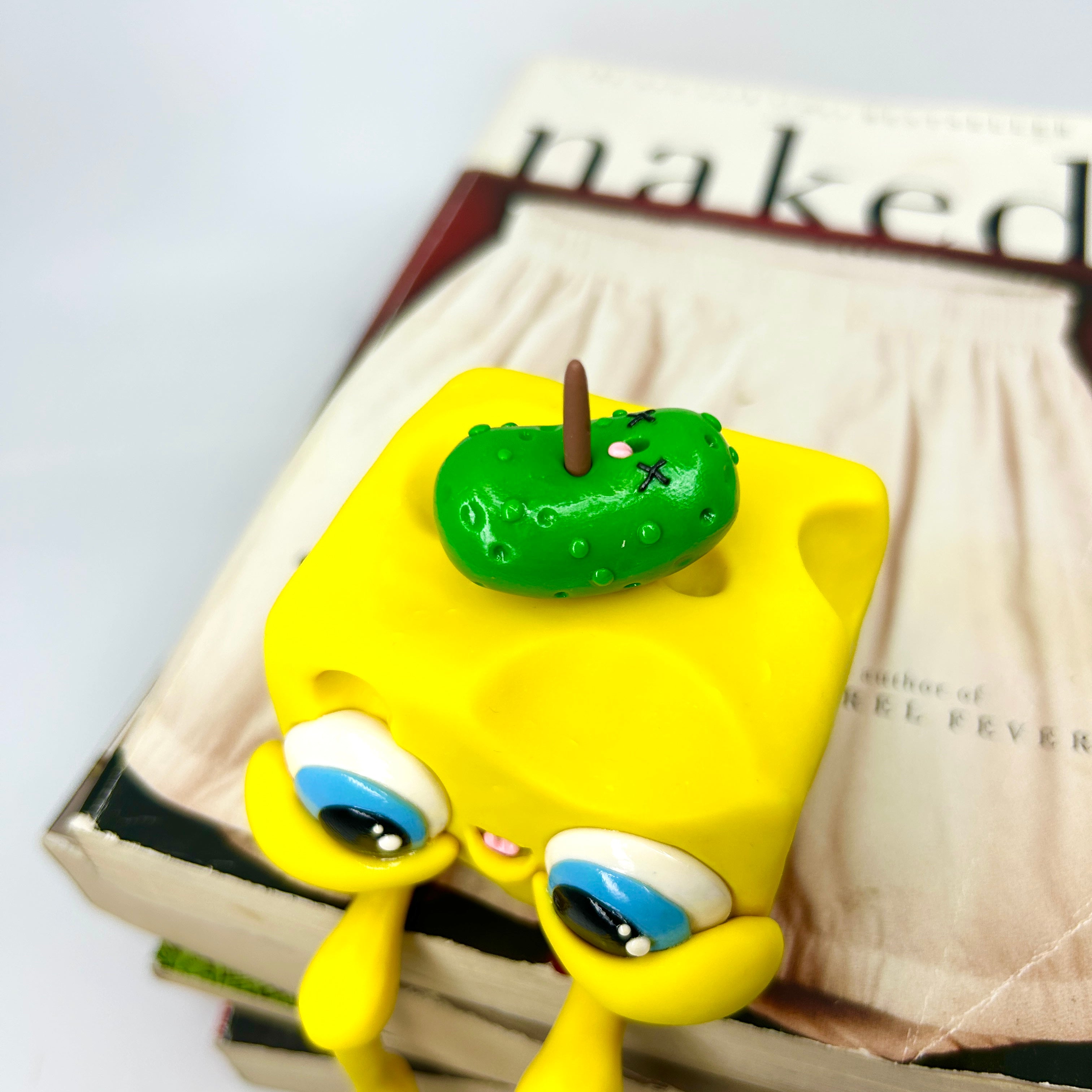 Toy cheese block with green pickle on books, close-ups of candy and toy eye from Simon Says Macy & Friends collection.