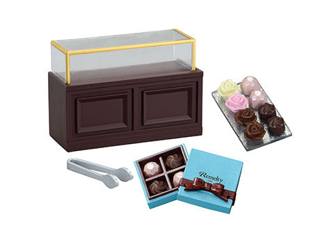 Miniature chocolaterie display from the Chocolaterie Re-ment Blind Box Series, featuring a counter, assorted truffles, tongs, and a branded gift box.