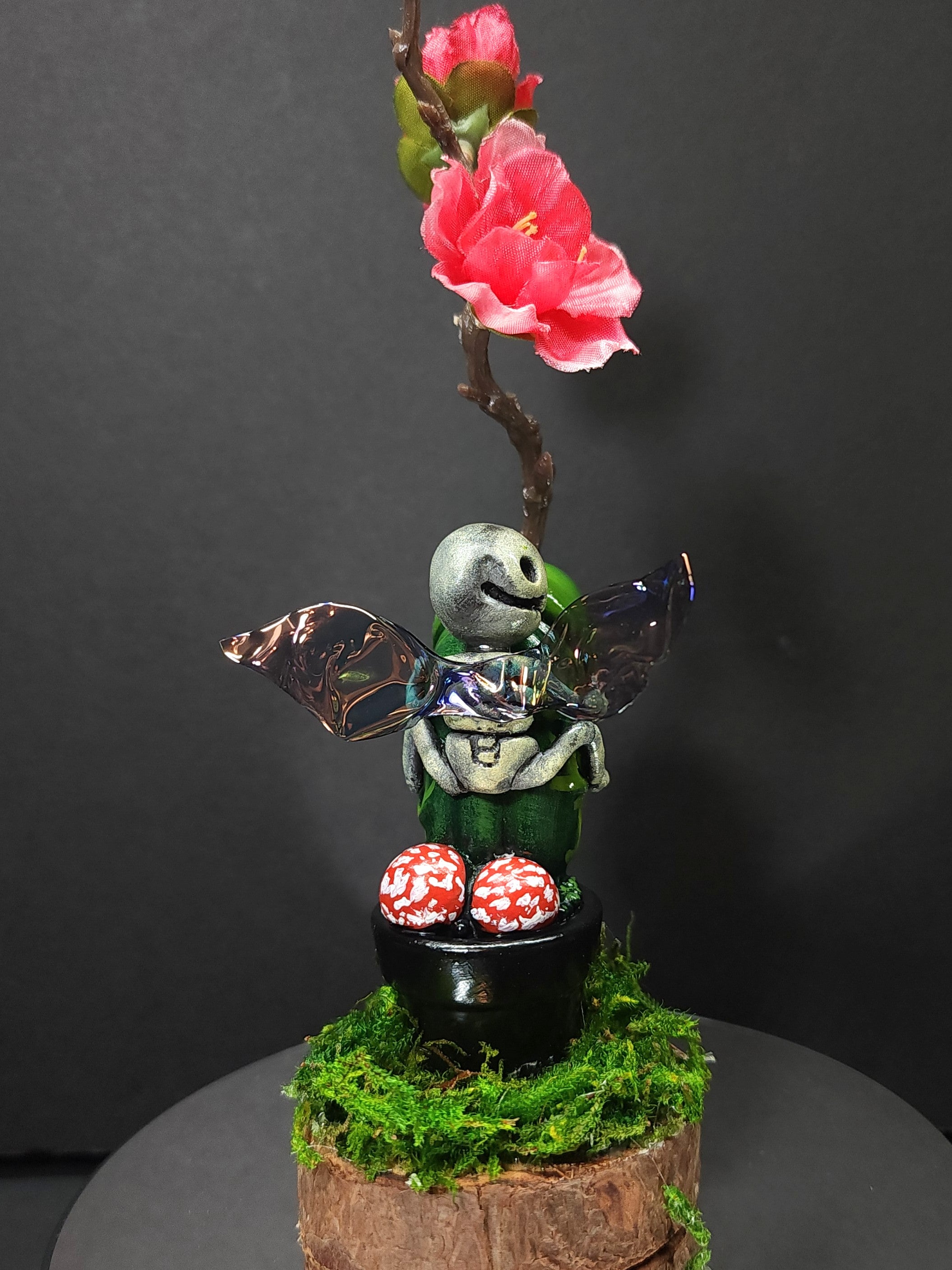Polymer clay statue of fairy on flowerpot with flower and mossy plant on stump.