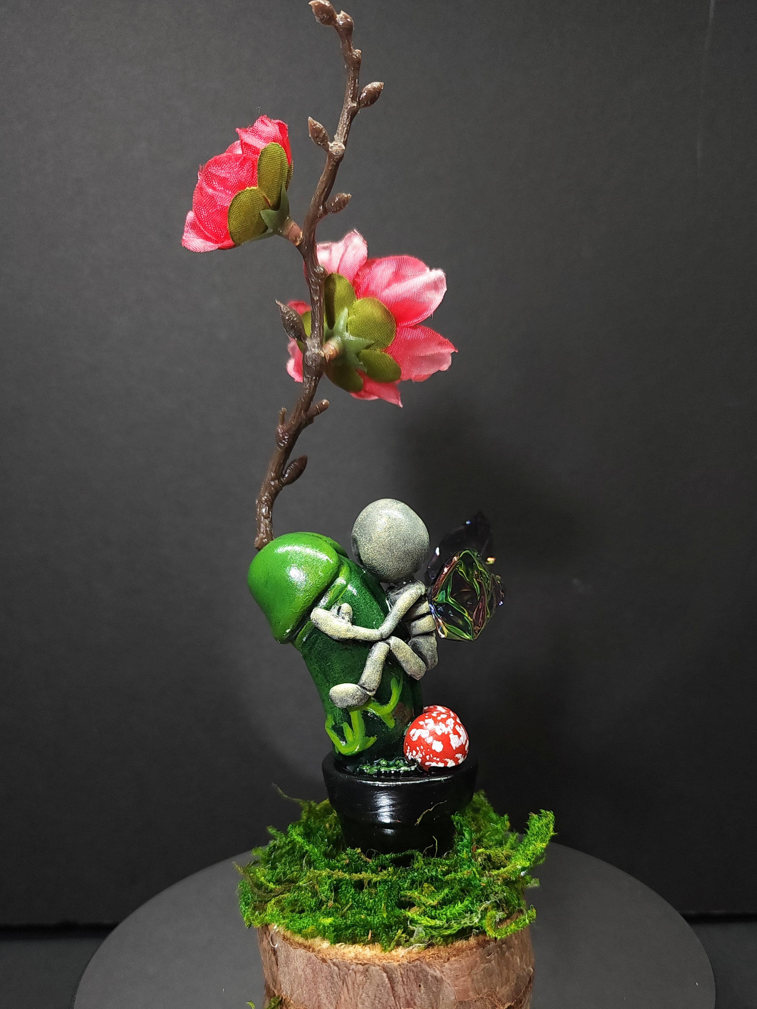 Polymer clay sculpture of a person holding a butterfly and flower, a mushroom, and a mossy stump.