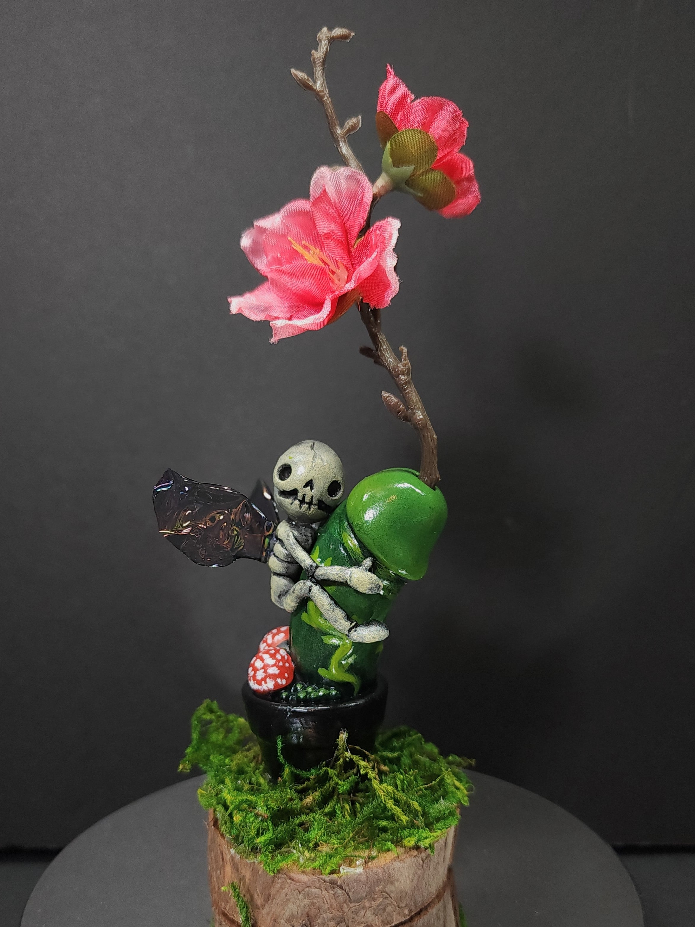A small statue of a skeleton holding a flower, a mossy tree stump, a toy figurine of a skeleton hugging a green pepper, a black object, a mushroom, a plant, a green object, and a flower.