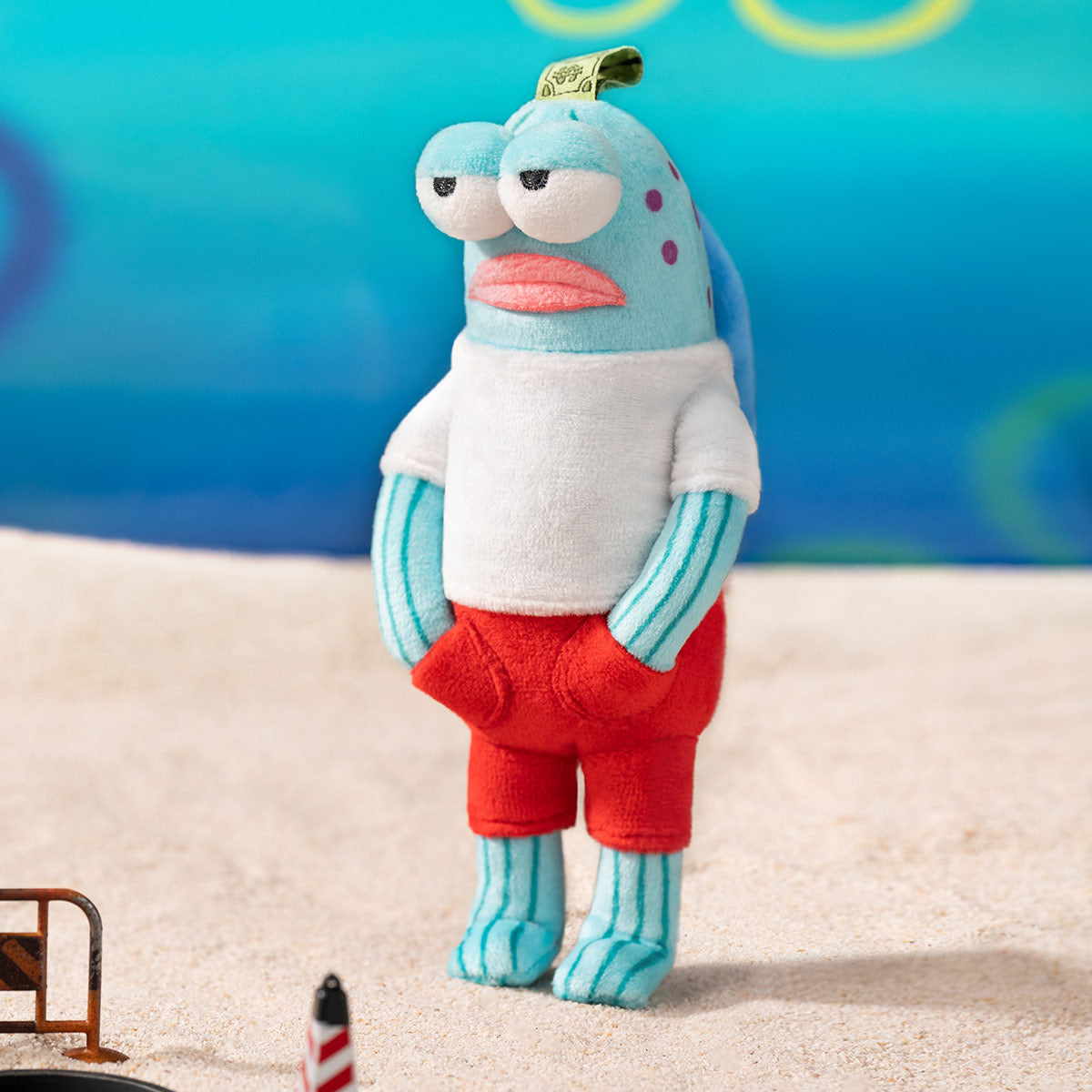 Bikini Bottom Buddies - Whimsical Plush: An aqua-blue fish-like toy in a white T-shirt and red shorts, standing on sand, ideal for playful collectors.