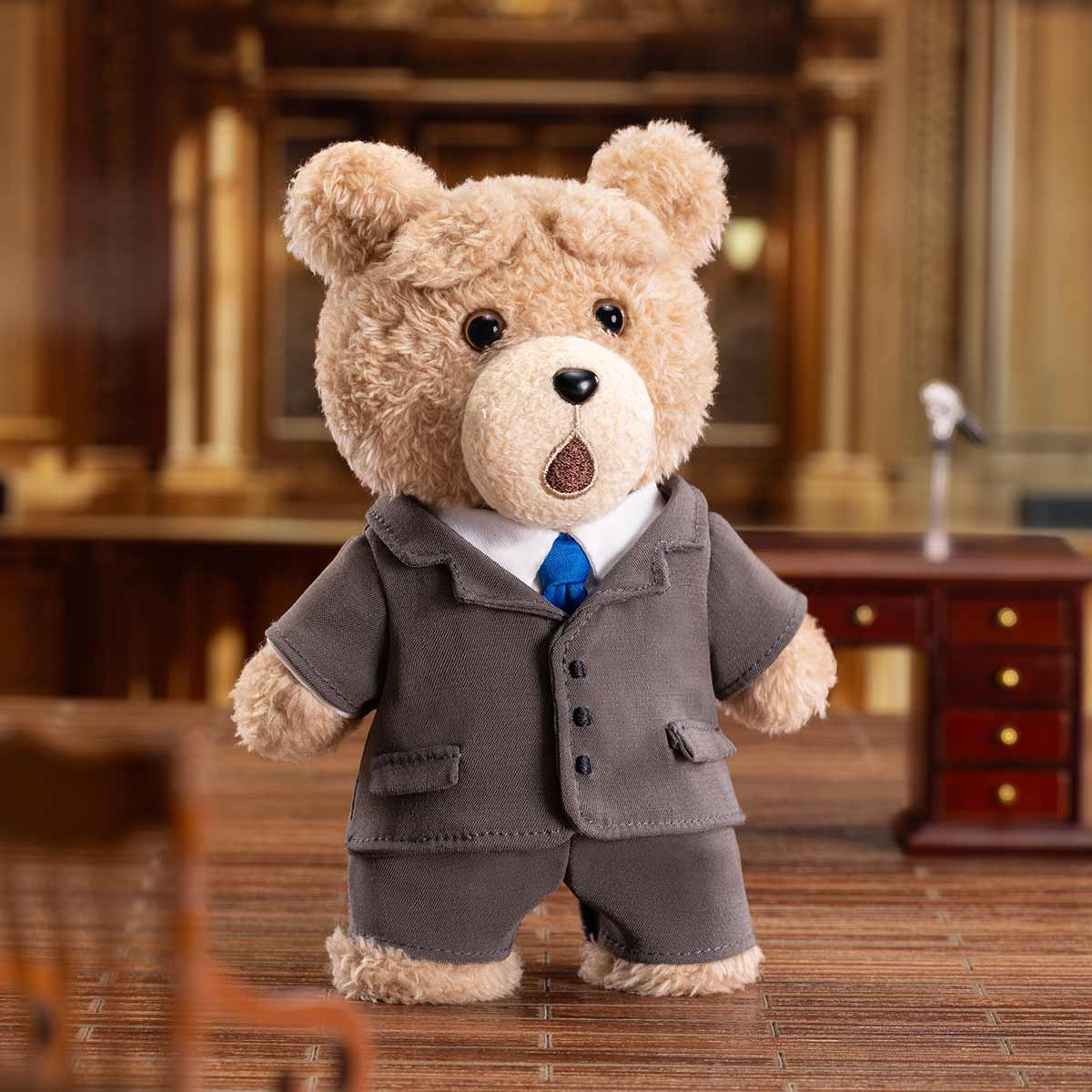 Ted2 Teddy Bear Action Plush Pendant in a gray suit and blue tie, standing on a wooden surface in a formal chamber setting.