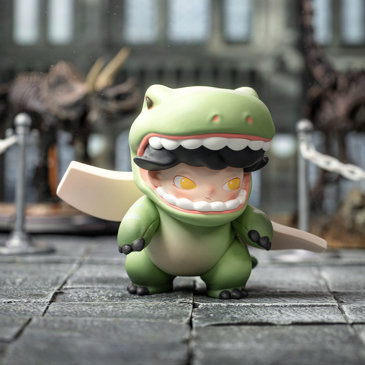 DIMOO Jurassic World Series figurine in a green T-rex costume, face visible, set in a museum with dinosaur skeletons. Preorder available, ships by Oct 2025.