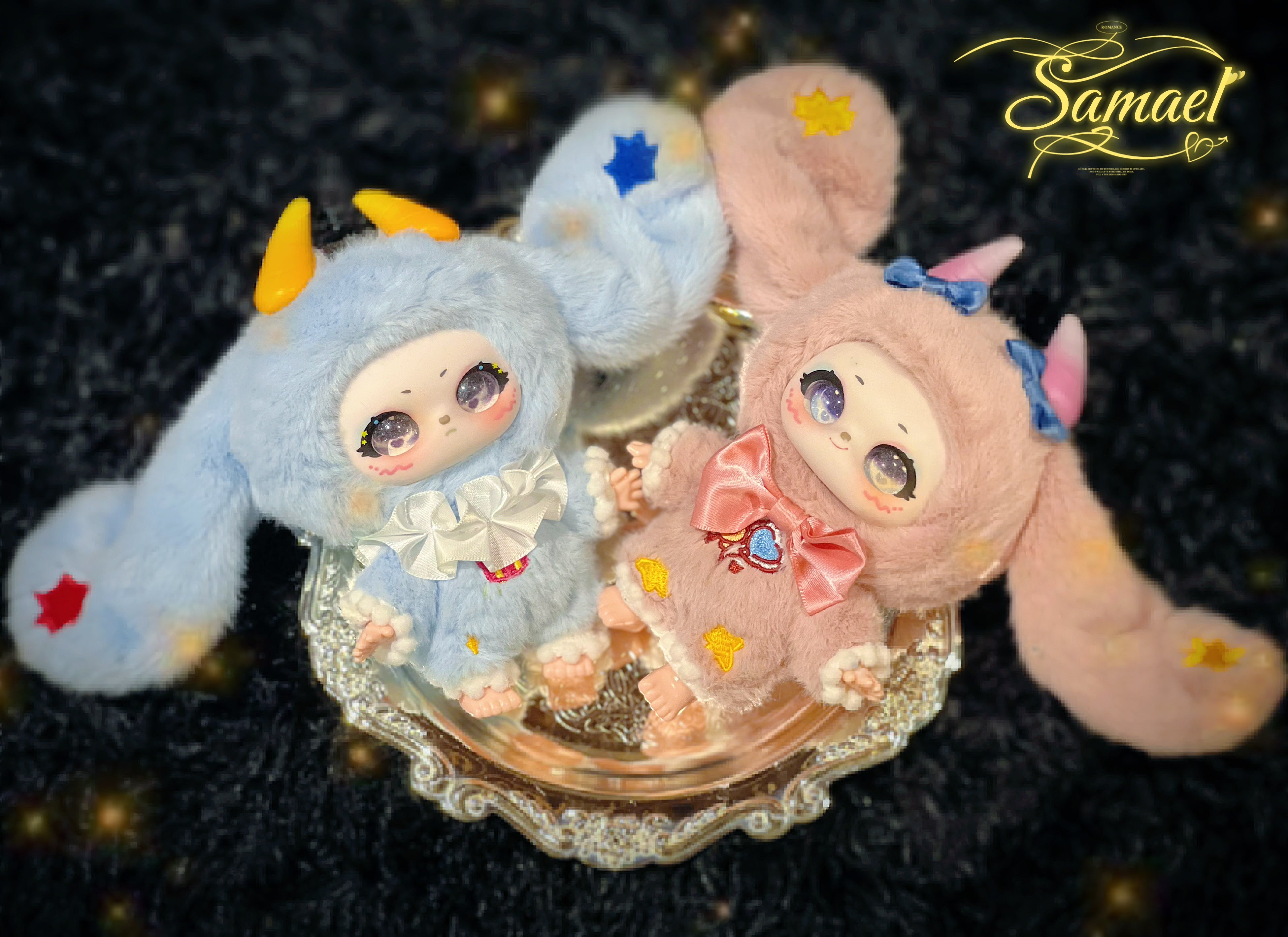 Two plush figures, Baby Three - Shamali Magic Wardrobe, in animal onesies, posed on a silver platter, featuring star appliqués and glittery eyes.