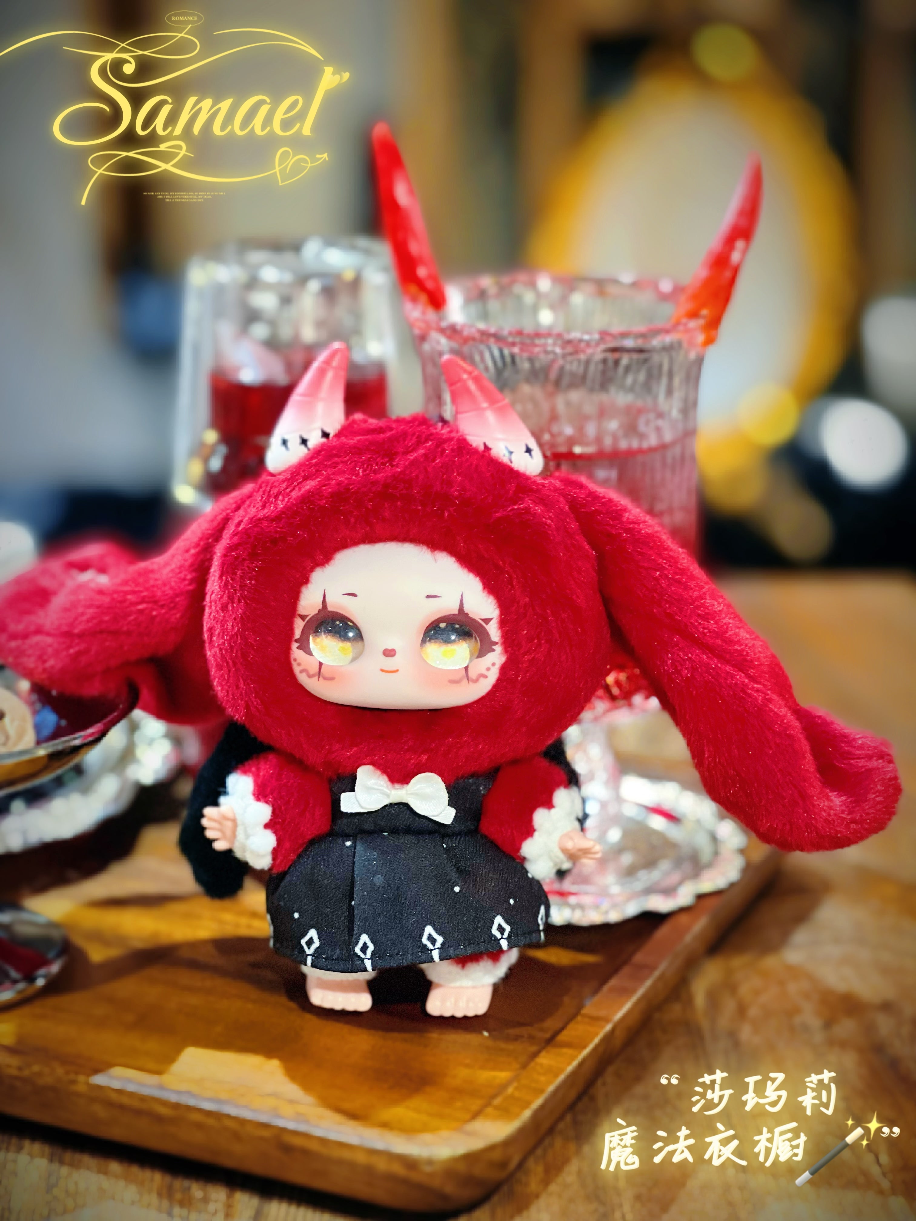 Baby Three figurine from Shamali Magic Wardrobe, featuring big eyes and a red hood with horns, seated on a tray with glassware in the background.