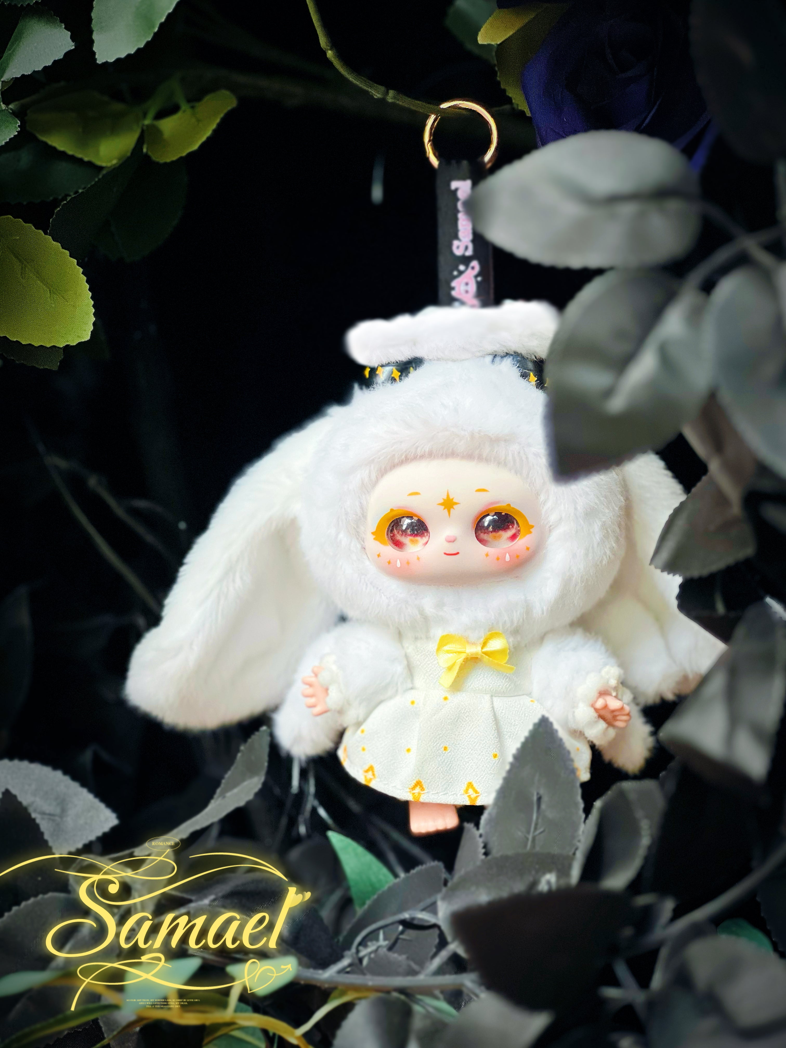 Baby Three - Shamali Magic Wardrobe plush keychain in bunny outfit with sparkly eyes, yellow bow, on gold ring, framed by foliage.