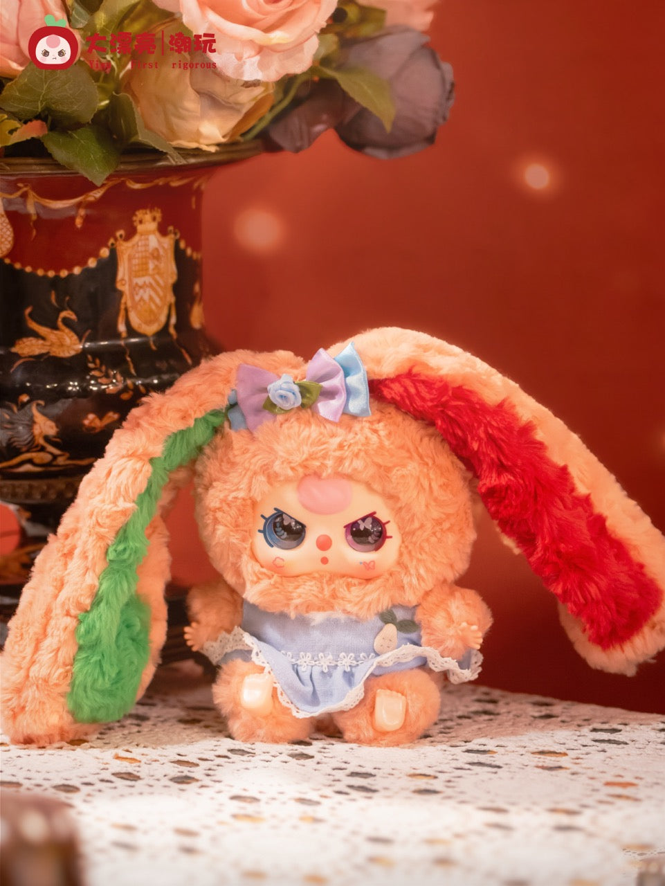 Baby Three - Lily Rabbit Town 4: A peach-orange plush rabbit in a blue pinafore, with colorful ears, sits on lace, alongside a decorative black-and-gold vase.