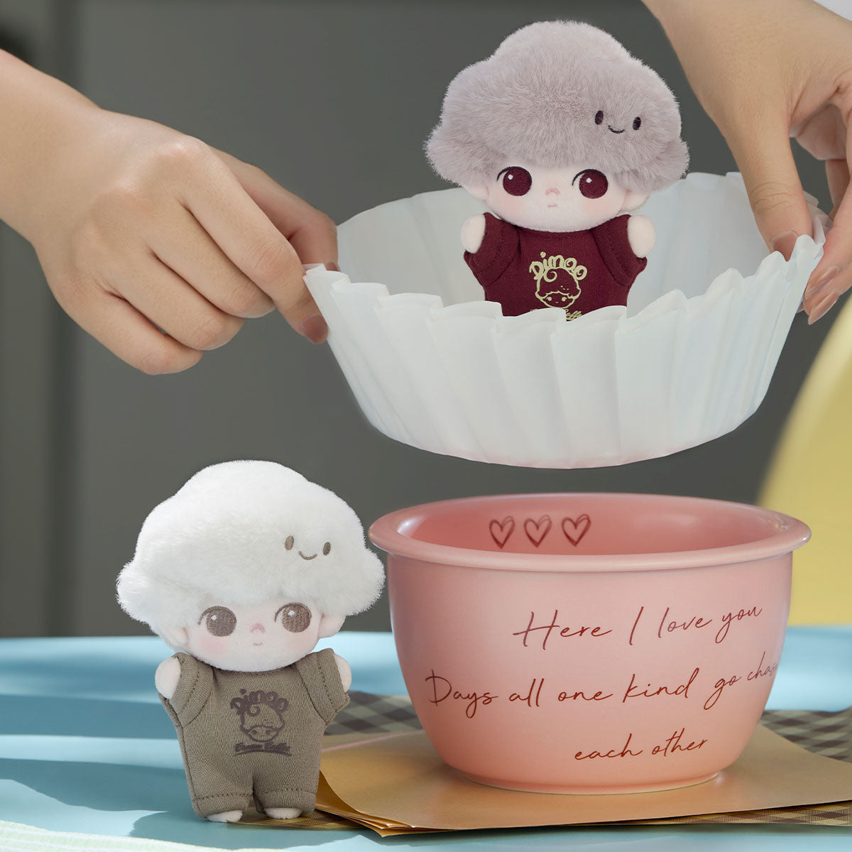 Two DIMOO Crush on Coffee Series plush keychain dolls beside a pink bowl, with hands holding a mold above it, showcasing a playful, collectible scene.