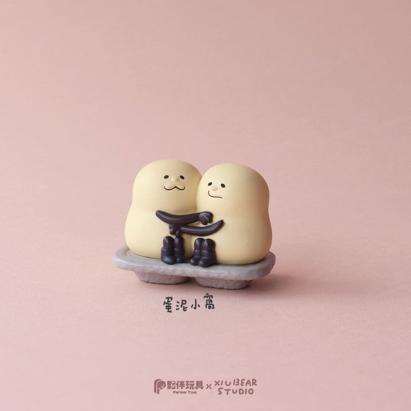 Yum Yum Time Blind Box Series: Egg-shaped figurines in an embrace on a gray stand, part of a designer toy collaboration.