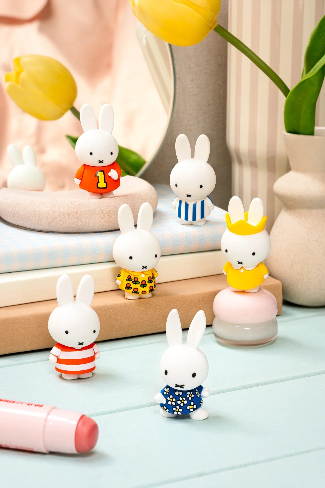 Smoko Miffy Dressing Up Blind Box Figure: Various Miffy figurines in playful outfits on pastel books, with a spring-themed setup featuring tulips and a makeup stick nearby.
