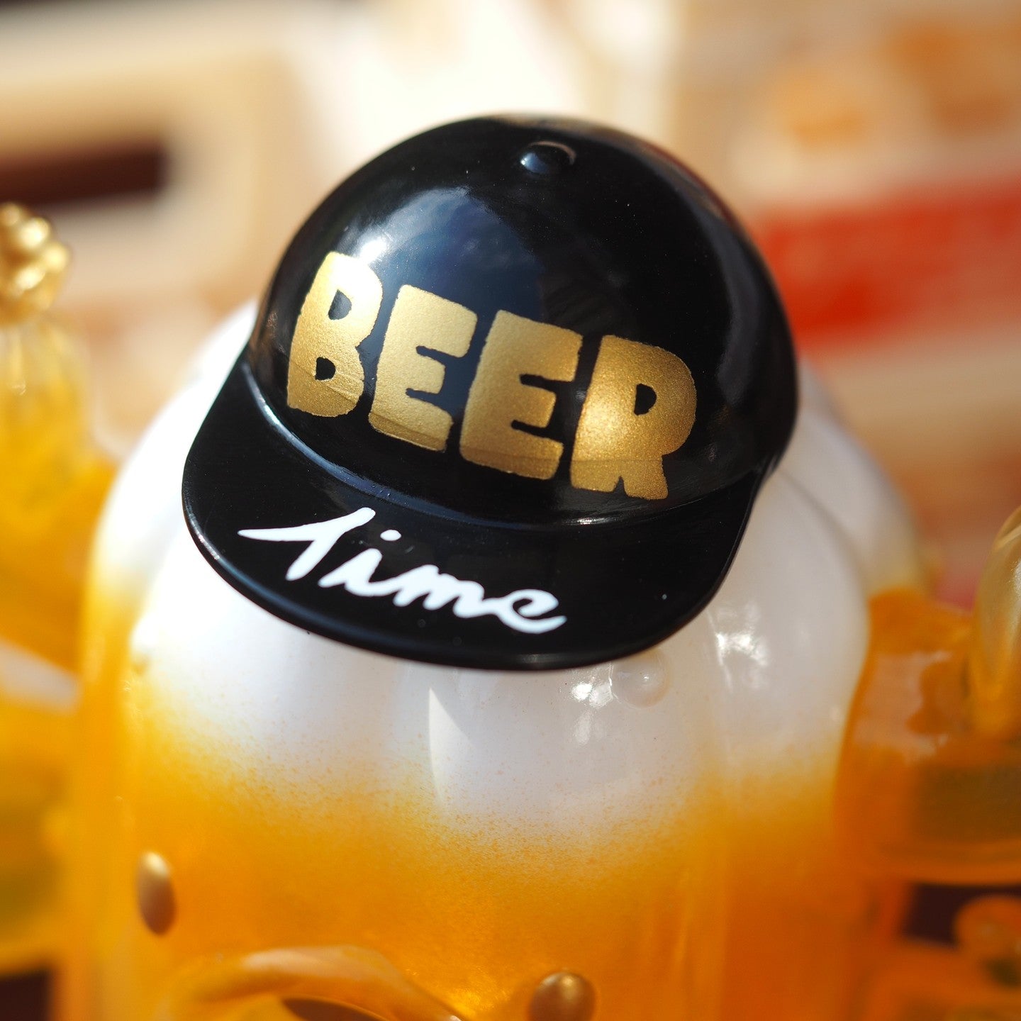 A black and white hat with gold text, a plastic toy resembling a hat, and a beer bottle in close-up. Product: BEER PATU, Soft Vinyl, 10 cm tall.