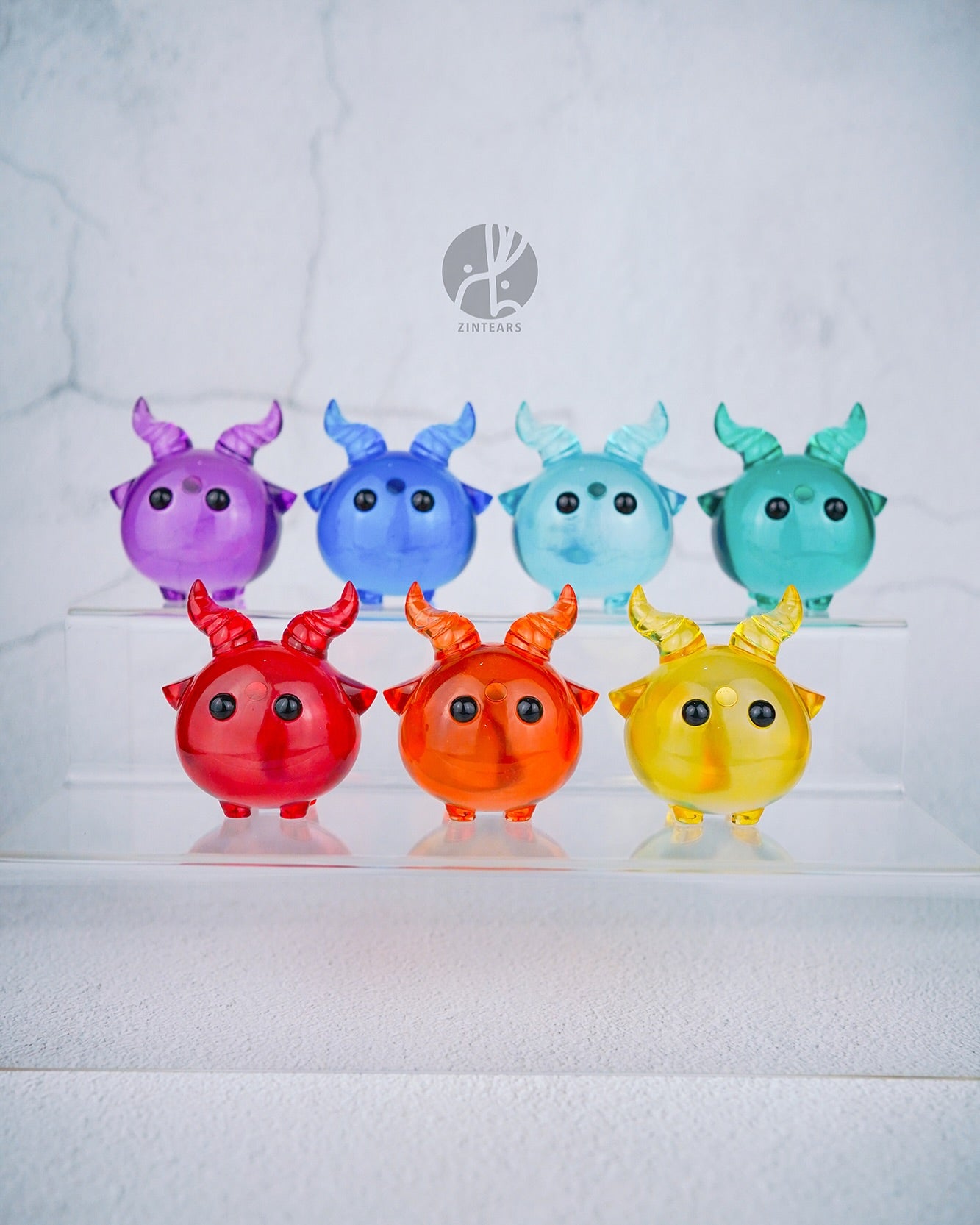 Yorick the skull - Orange Edition by Zintears, a small, horned resin figurine, stands prominently among rainbow-toned collectibles on clear acrylic steps.