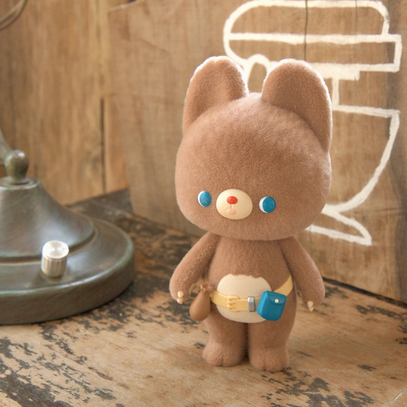 KUMARU THE FOREST BEAR FOREST FIXES - Preorder; a small plush figure with blue eyes and a belt, standing on wood near a lamp.
