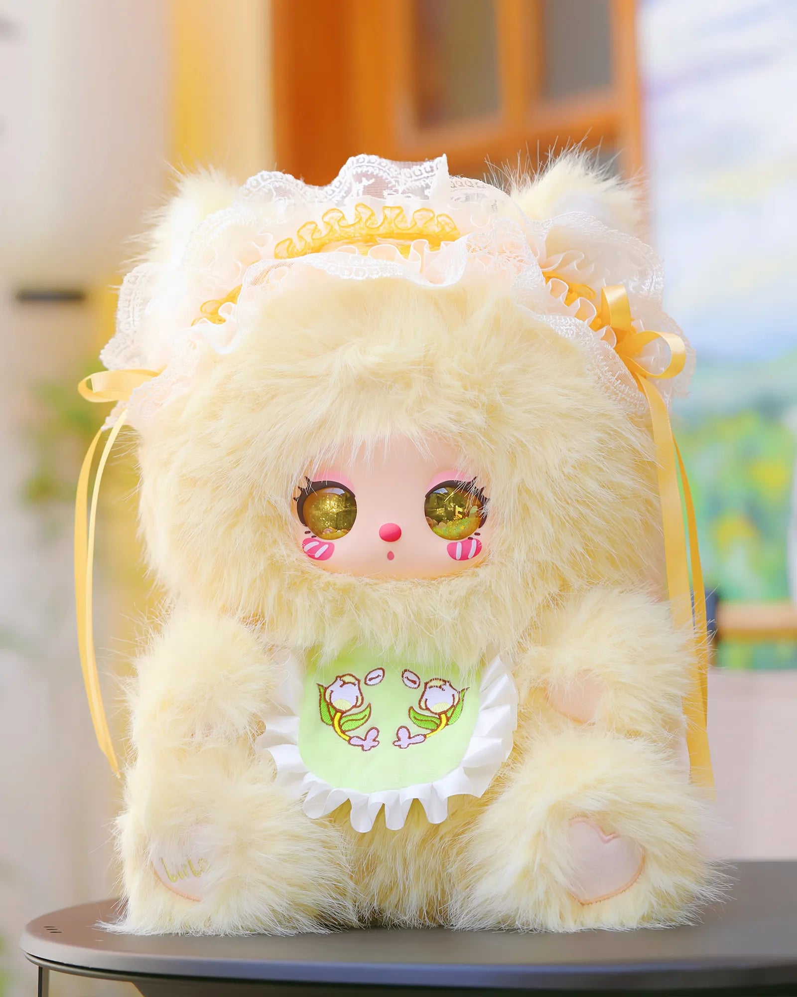 Liila's 600% 2V Tea Time With My Girl Plush features a buttercream faux fur doll with glittery eyes, bonnet, and tulip-appliquéd bib, on a round table.