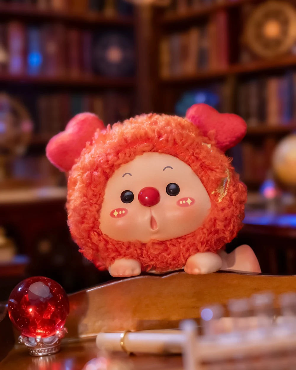 OYO Constellation Story Hipper Series Mini Blind Box figurine in an orange onesie with heart ears, placed on a desk near a red orb and pen.