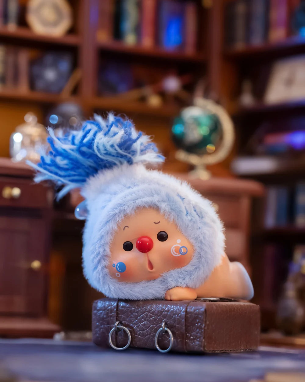 OYO Constellation Story Hipper Series Mini Blind Box toy, a rosy-faced figurine with a fluffy blue hood, on a leather-textured box in a cozy setting.