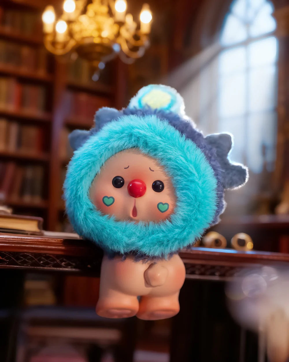 OYO Constellation Story Hipper Series Mini Blind Box toy with a turquoise hood, cartoon face, on a wooden table in a book-lined study.