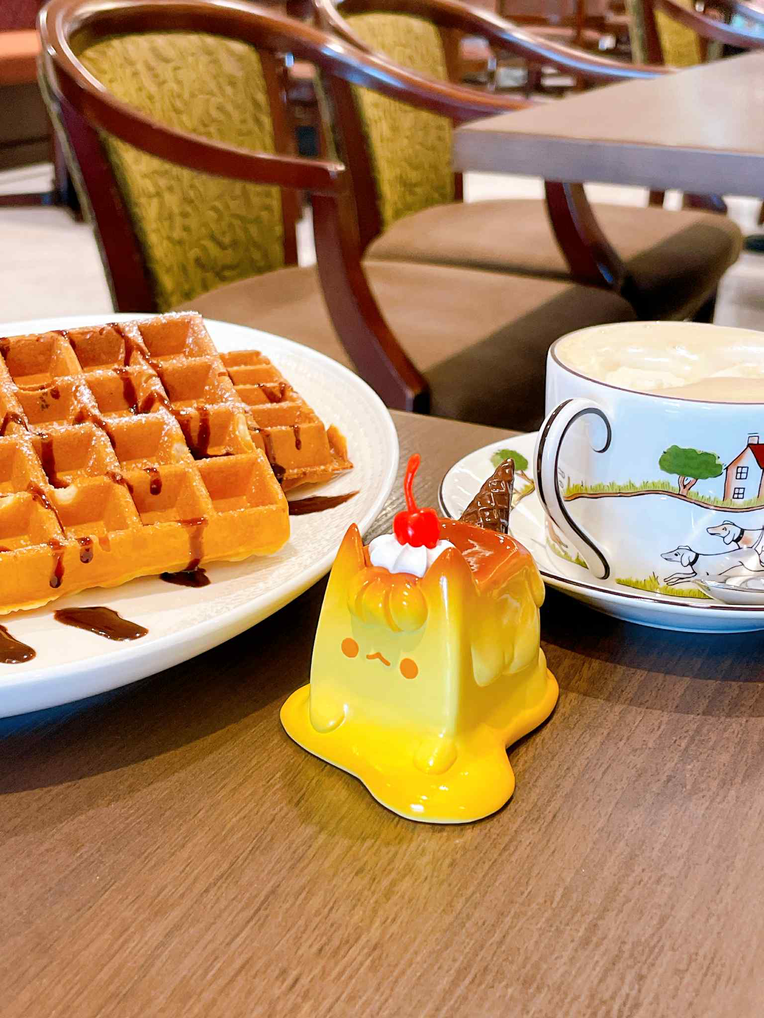 Pudding Cat by Rato Kim, a whimsical cat-shaped toy resembling a dessert with whipped cream, cherry, and mini waffle cone, atop a cafe-style setting.