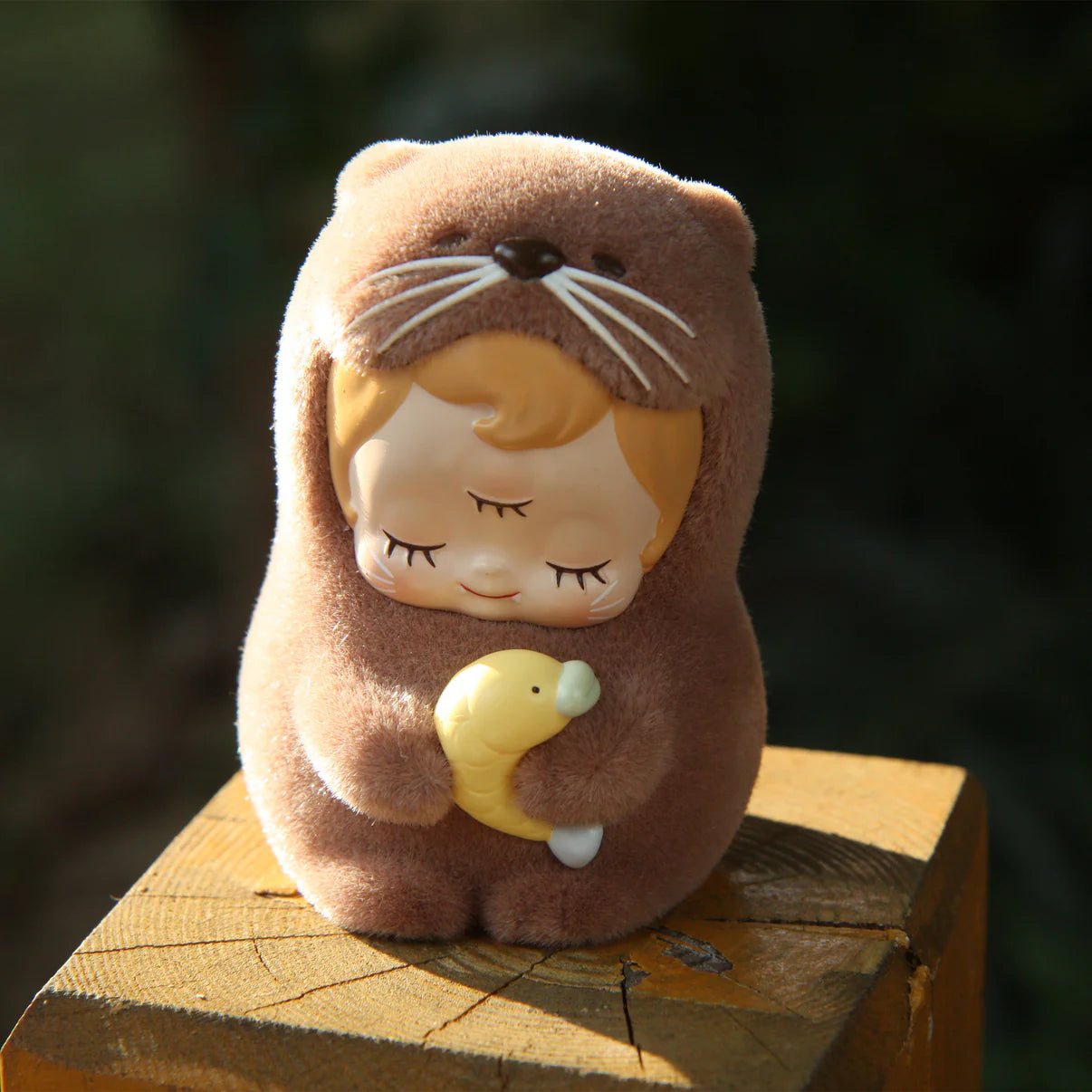 ABAO-Otter baby dede figurine in otter costume with closed eyes, holding a fish. Perched on a wooden block, available for preorder at Strangecat Toys.