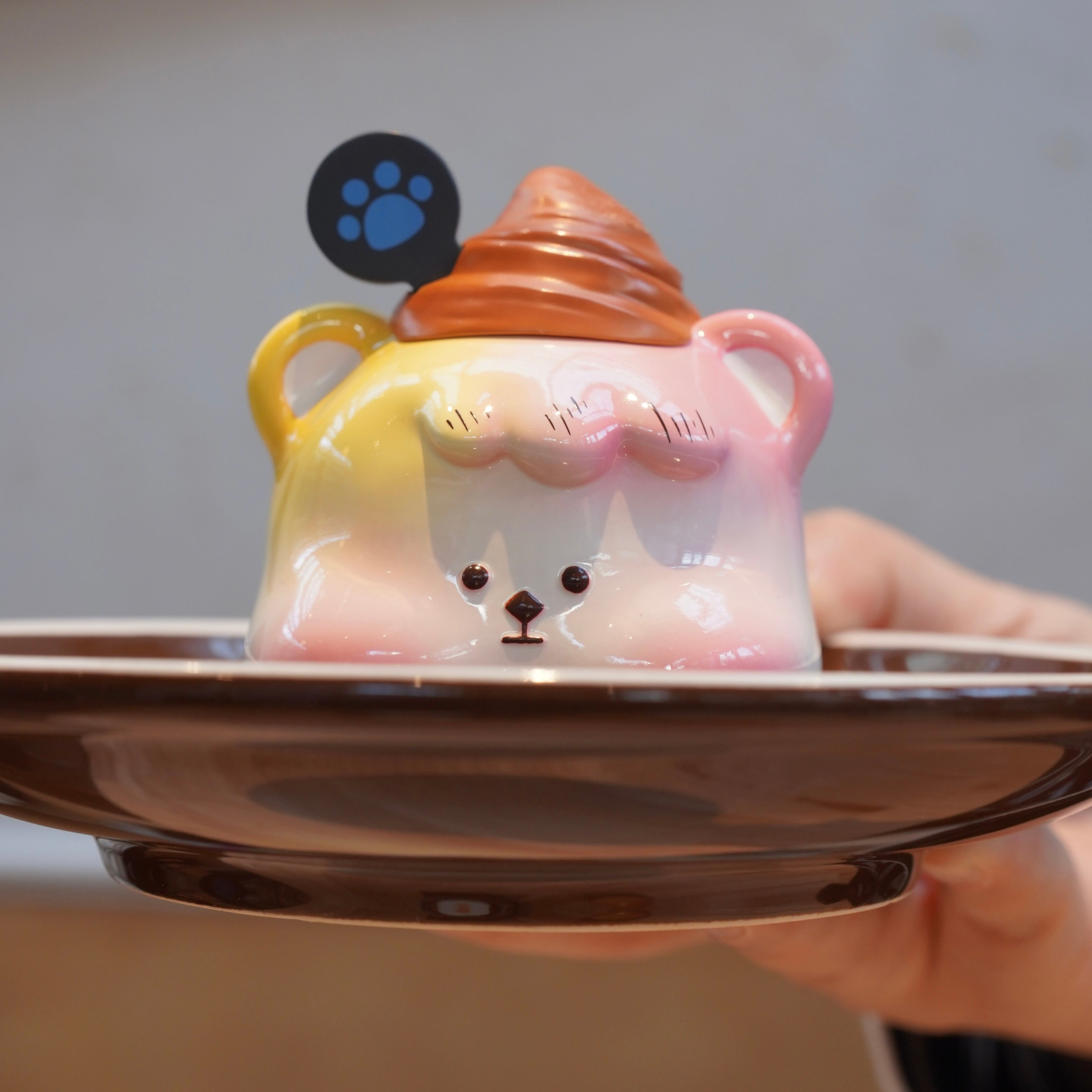 A hand holding a plate with a small toy, a ceramic bear, and a blue paw print, part of Croissant Han Han by Unknown Island - Preorder.