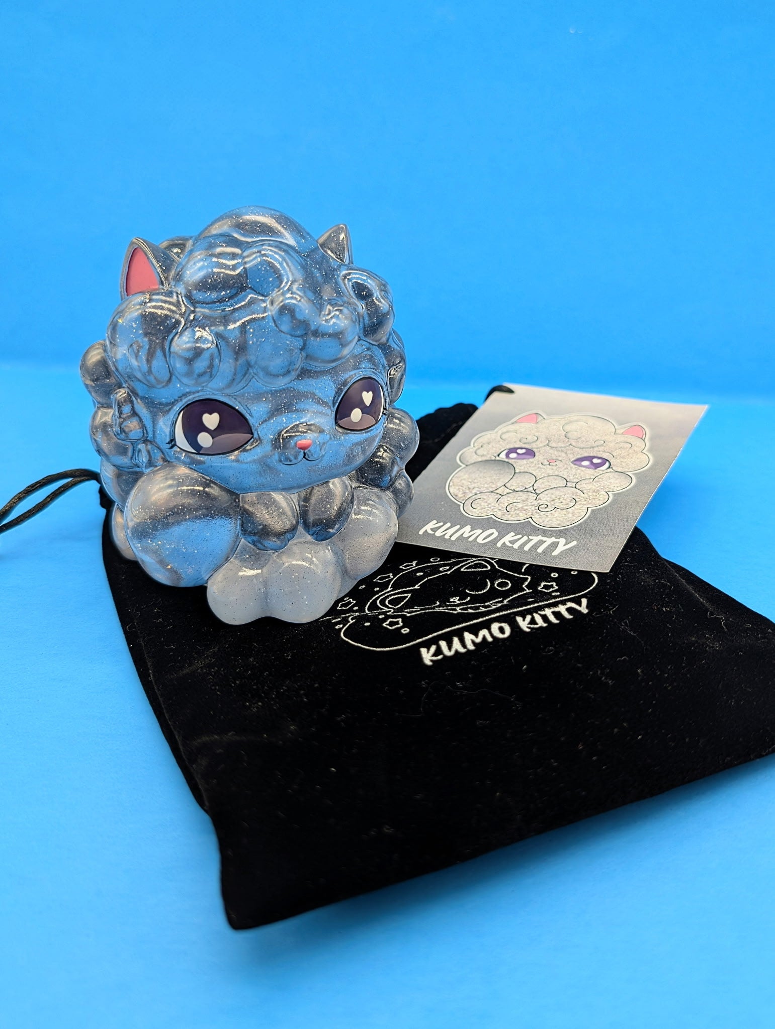 Kumo Kitty - Stormy Skies figurine with glittery, cloud-like fur, sits on a Kumo Kitty pouch, accompanied by an art card showing the same design.