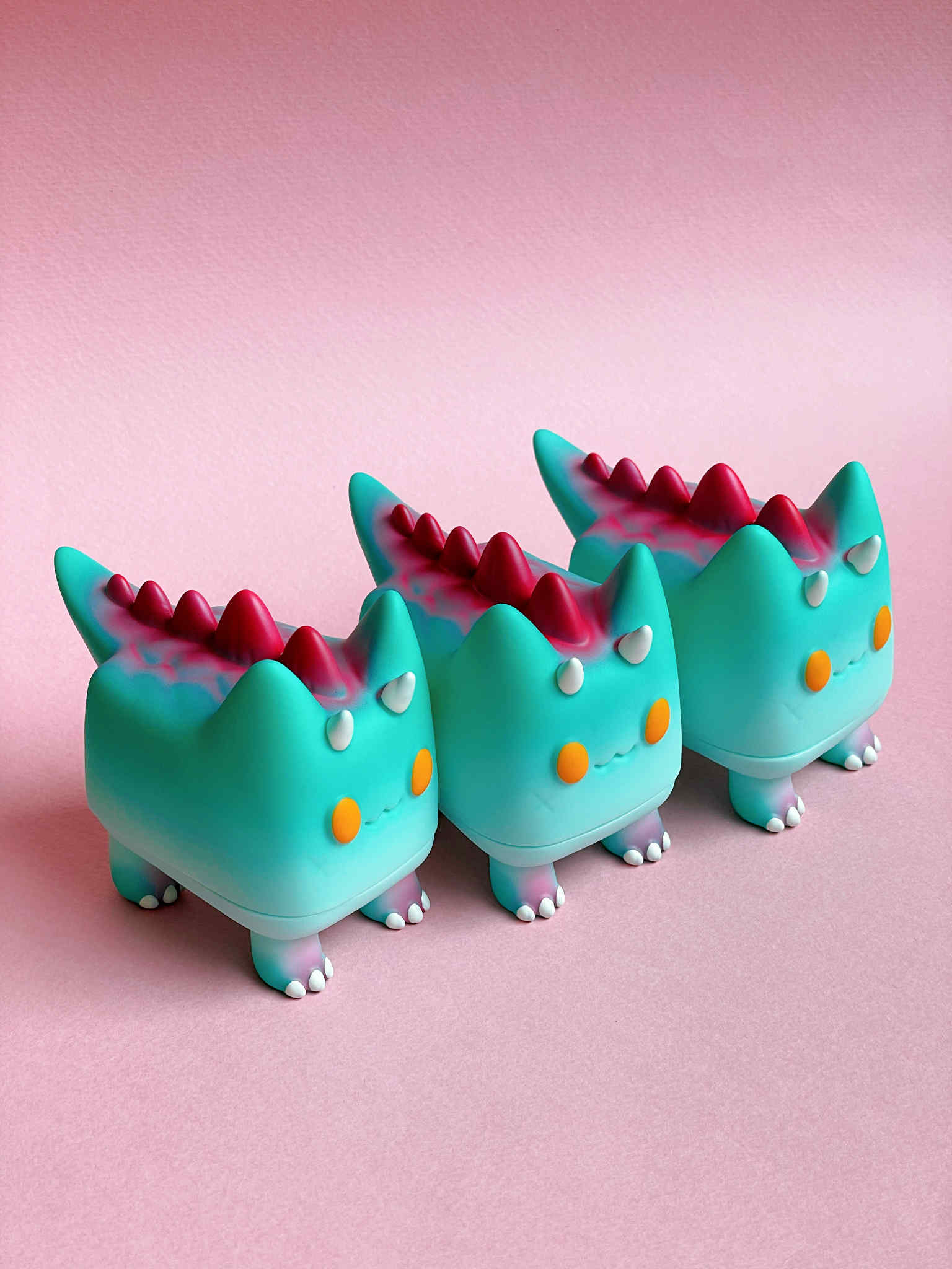 DiDi (Dino cat) by Rato Kim - Preorder: Three identical squat figurines with horns and spikes, crafted in sofubi, lined up on a pale pink surface.
