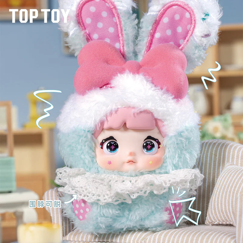 Nommi Baby Sweetheart Bunny Plush Blind Box doll in a bunny costume with sparkly eyes, pink hair, and a pink bow, on a striped cushion.