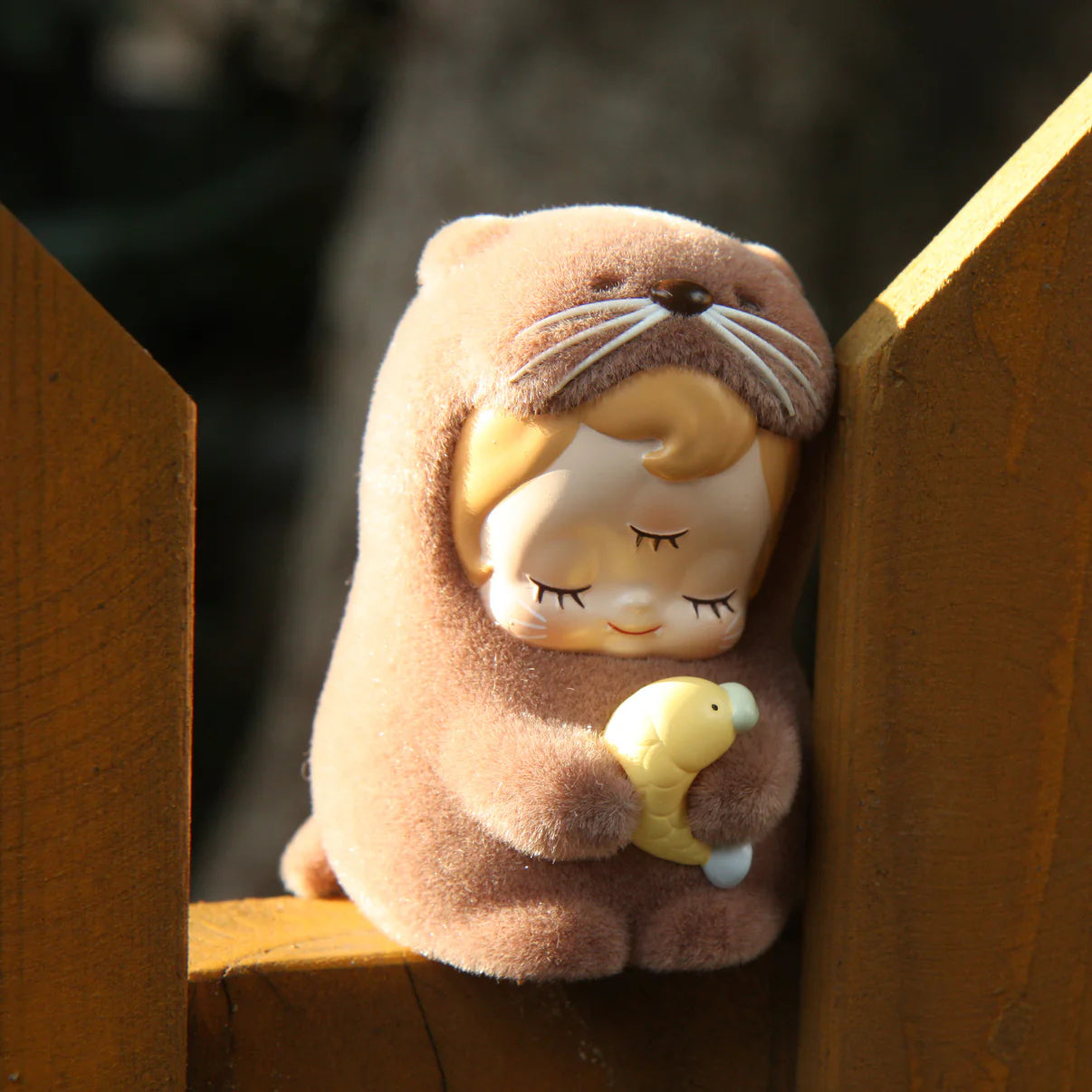 ABAO-Otter baby dede - Preorder: A 7.5cm plush-covered resin figurine of a child in an otter costume, clutching a toy fish, on a wooden railing.
