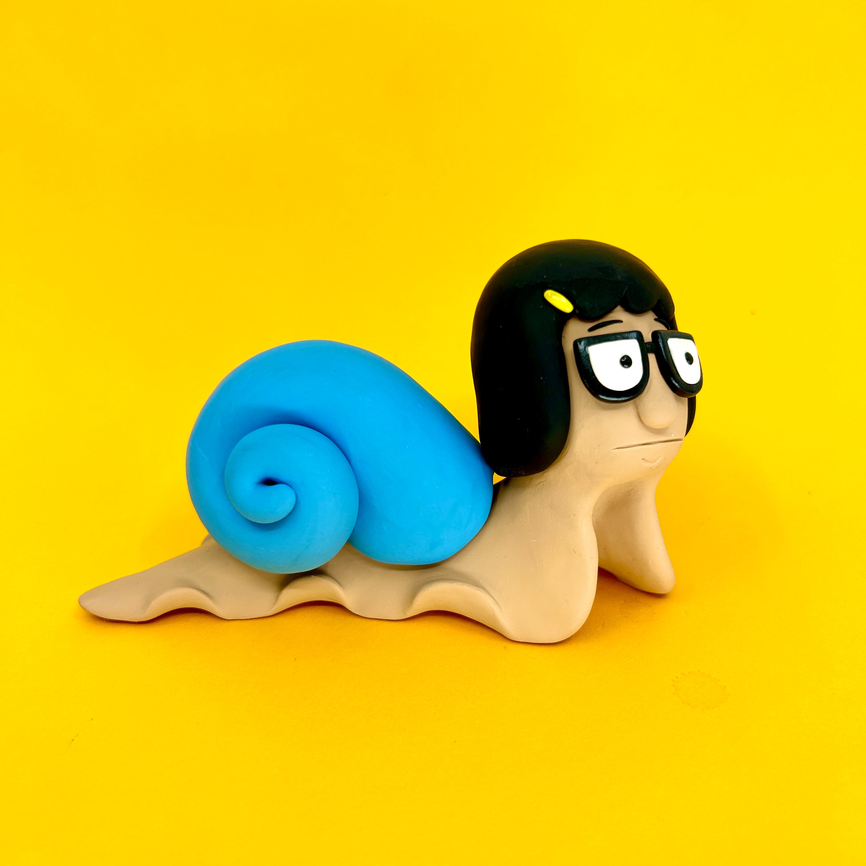 A clay snail with glasses and a blue shell, part of Simon Says Macy & Friends - Tina Snailcher collection.