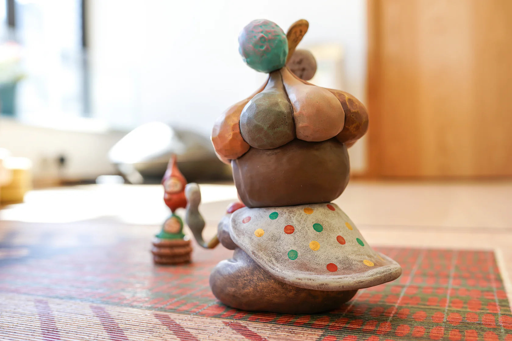 The Beanie Charmer - X-Mas Edition by Pucky, a hand-made ceramic figurine with a polka-dotted cape, seated on a mat, accompanied by tiny clay objects.
