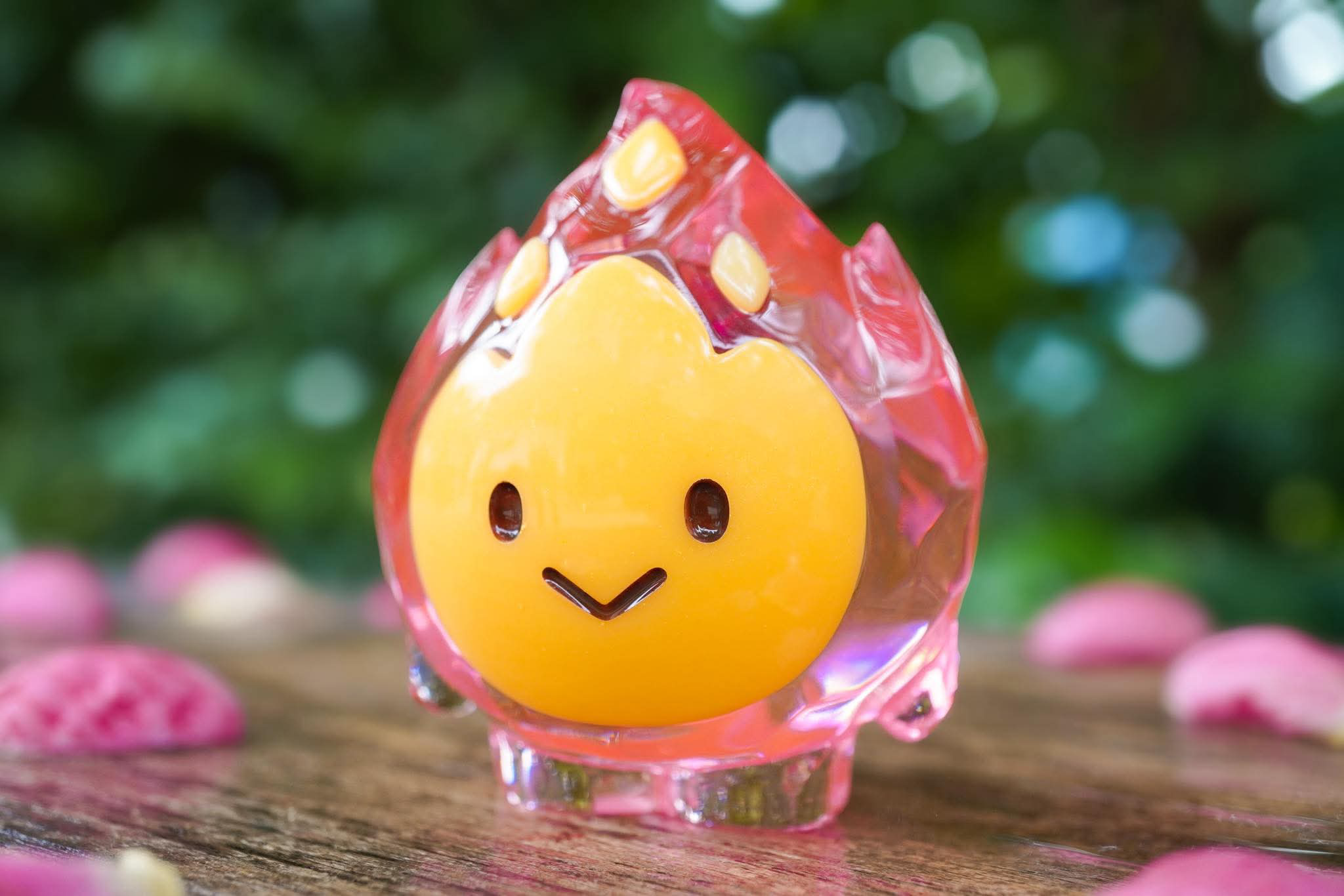 FLAMEY FROST by MUPA Toy, a 7cm translucent pink resin figurine with an orange smiling face, set among pink petals on a wooden surface.