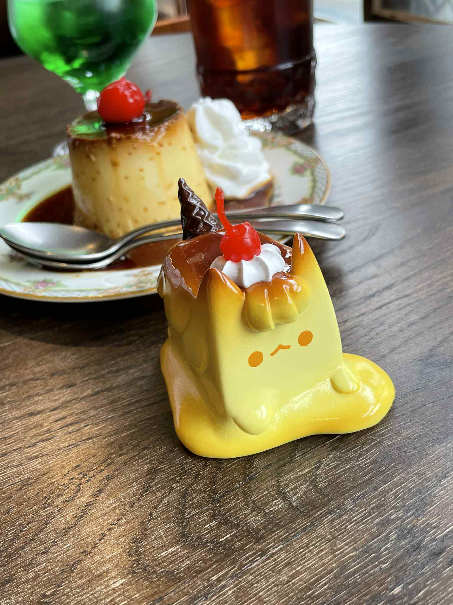 Pudding Cat by Rato Kim figurine with caramel top, cream, and cherry, displayed with real flan, green soda float, and iced drink.