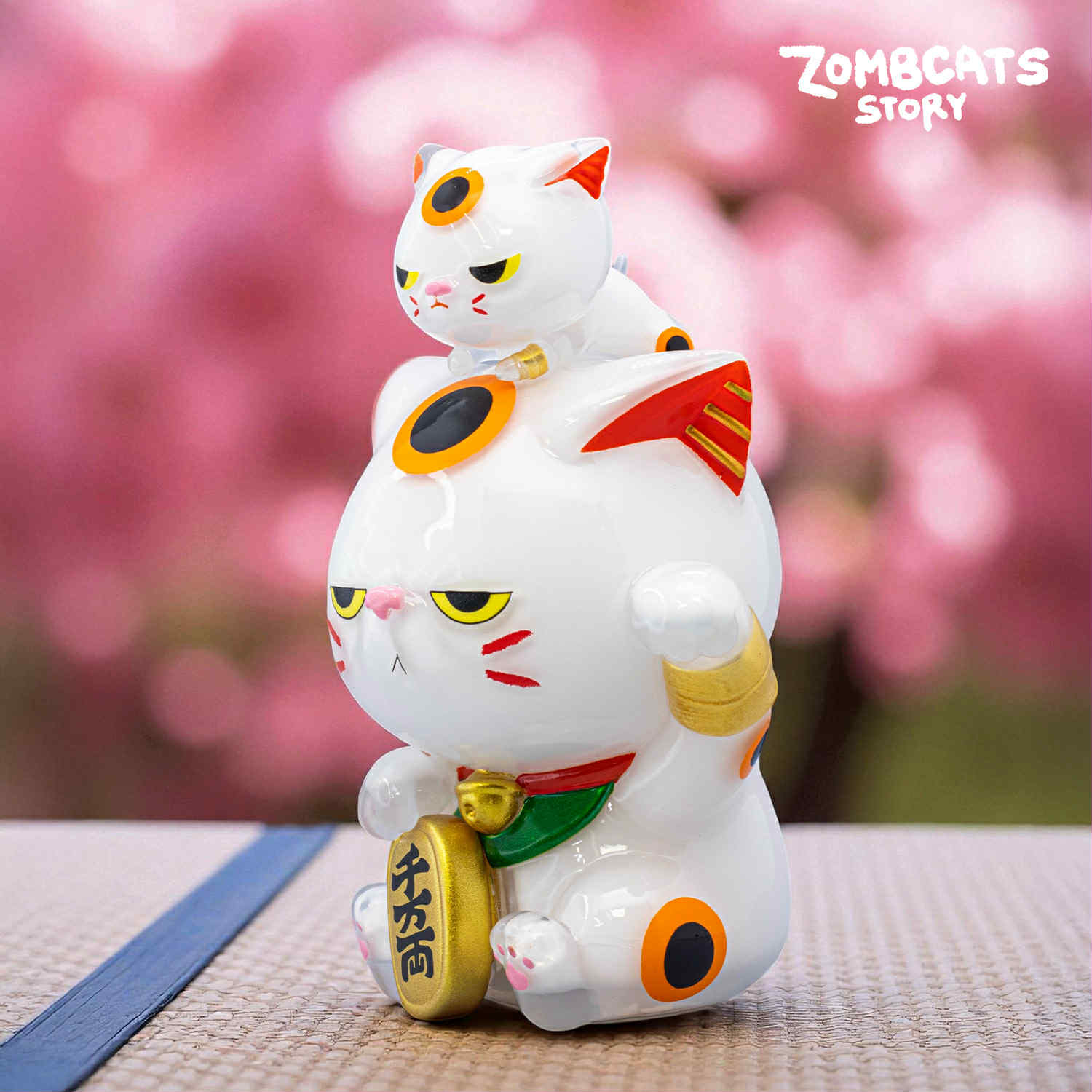 Zombcats Story - Grumpy Maneki LOTTERY figurine: a white lucky cat with red accents, holding a coin and topped by a smaller cat, set against a floral backdrop.