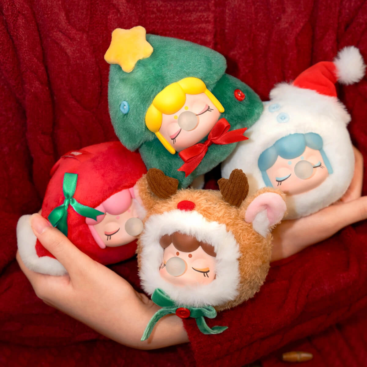Hands in a red sweater hold four Rolife Nanci's Christmas Gift Plush Pendants, each in unique holiday-themed costumes: tree, Santa, reindeer, and snowman.