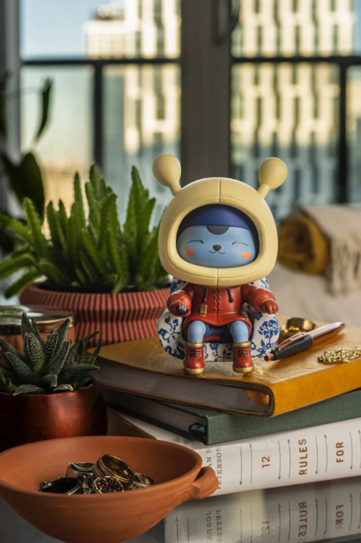 Napa - Porcelain: A whimsical alien figurine in red, seated on a patterned armchair atop books, surrounded by potted succulents and trinkets.