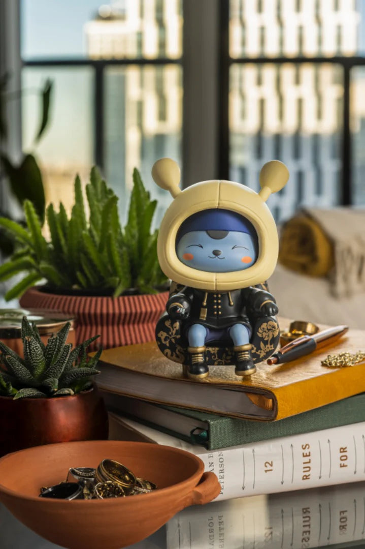 Collectible figure Napa - Obsidian, a blue character in a yellow hood, sits on a black-and-gold chair atop books, surrounded by plants and trinkets.