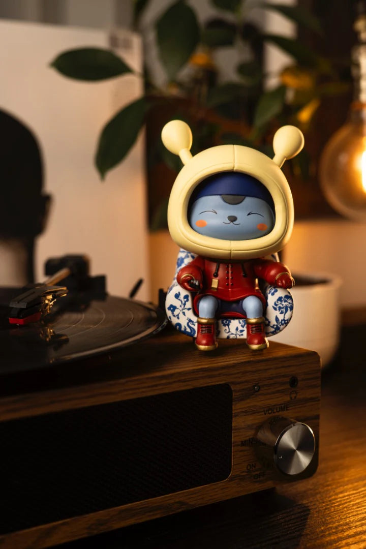 Napa - Porcelain Edition: A blue, cat-like vinyl figure in a red outfit, seated on a floral-patterned chair atop a turntable, embodying limited-edition art toy charm.