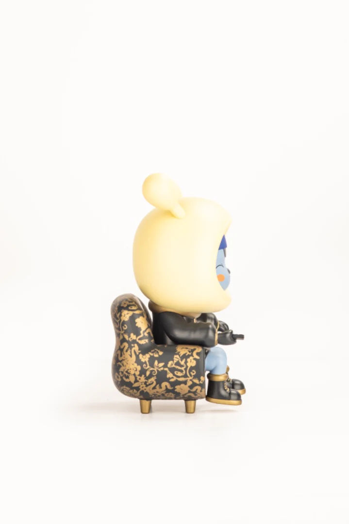 Napa - Obsidian vinyl figure in a gold-accented chair, wearing a yellow hooded cap, holding a remote, embodying alien exploration of human media.
