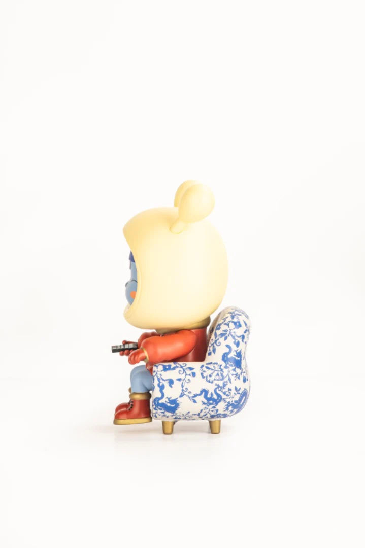 Napa - Porcelain figurine, an art-toy with a yellow head and red outfit, sits on a patterned chair, holding a small controller.