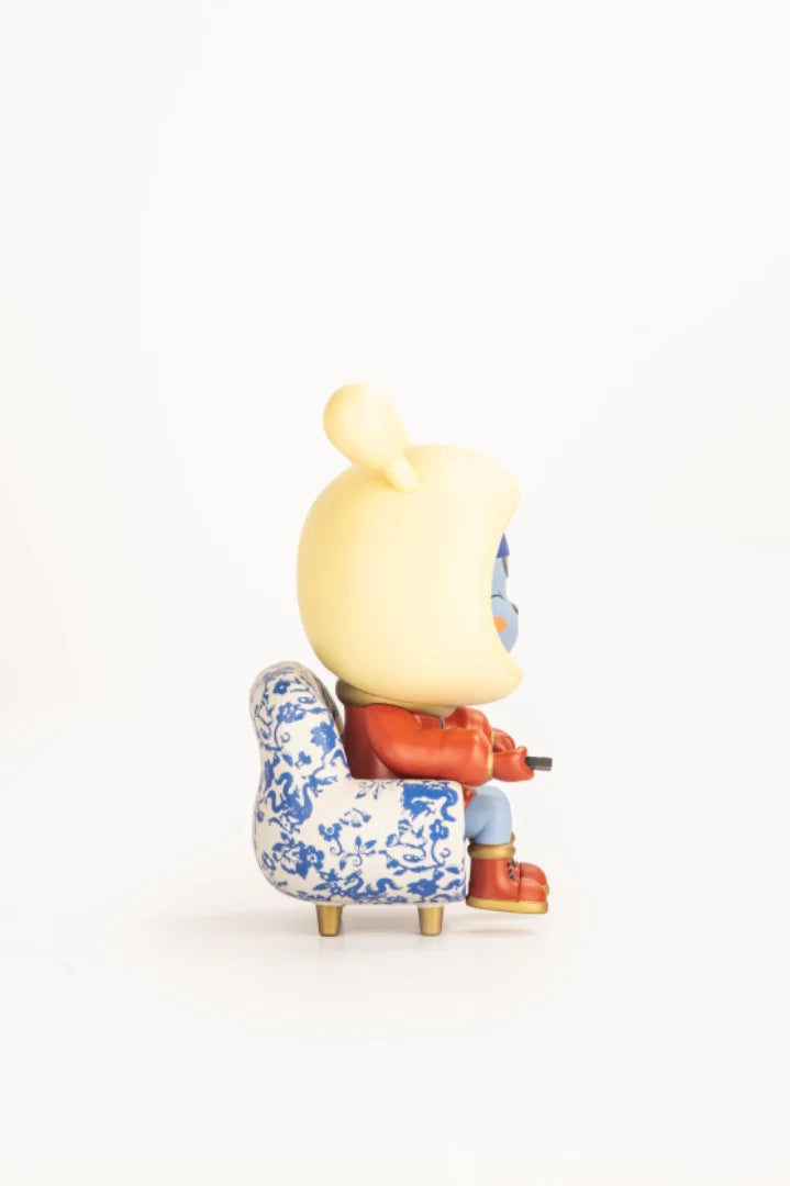 Napa - Porcelain art toy seated in a blue-and-white patterned armchair, holding a rectangular object, wearing a yellow hood and red jacket. Limited edition collectible.